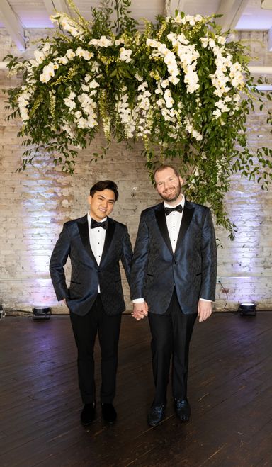 Two Grooms pose for a picture under elaborate hanging florals